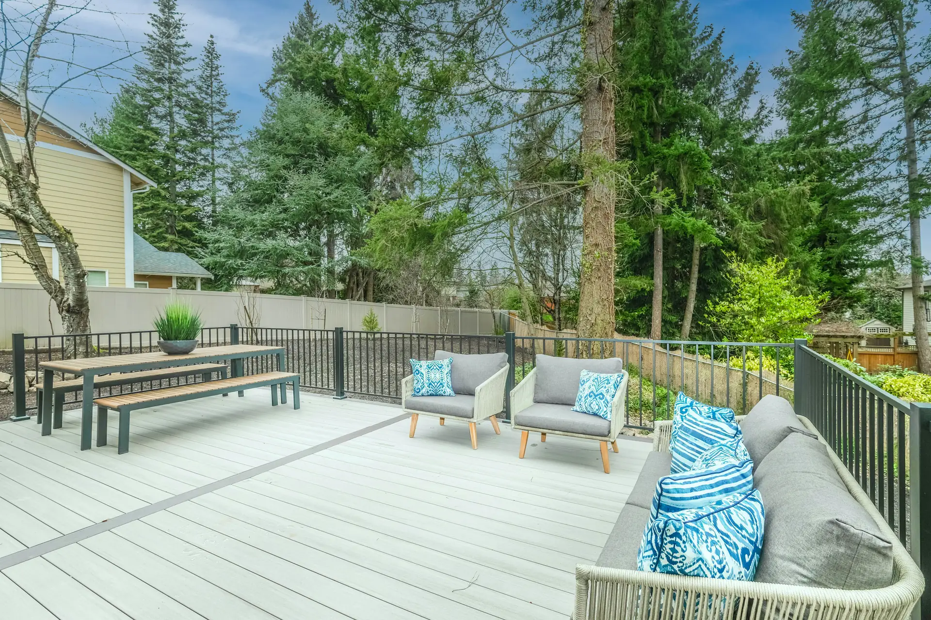 Stock photo: a light grey composite deck with a darker border board, black metal railing and outdoor furniture, backed by evergreen trees