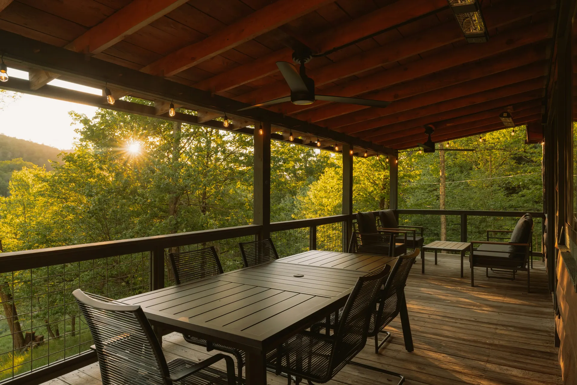 Stock photo: a covered wood deck under a timber-framed roof with ceiling fans and string lights, a dining table and a wire-panel railing